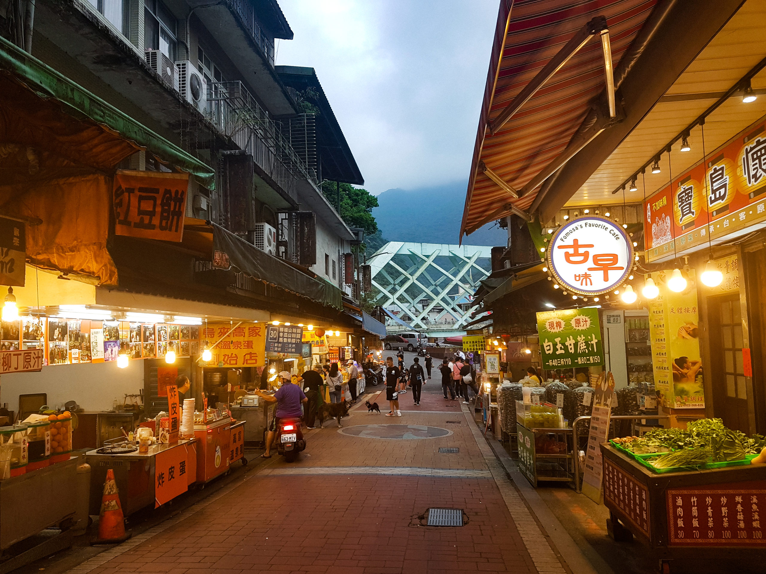 A bustling Asian street market at dusk, lit by warm orange street lighting.