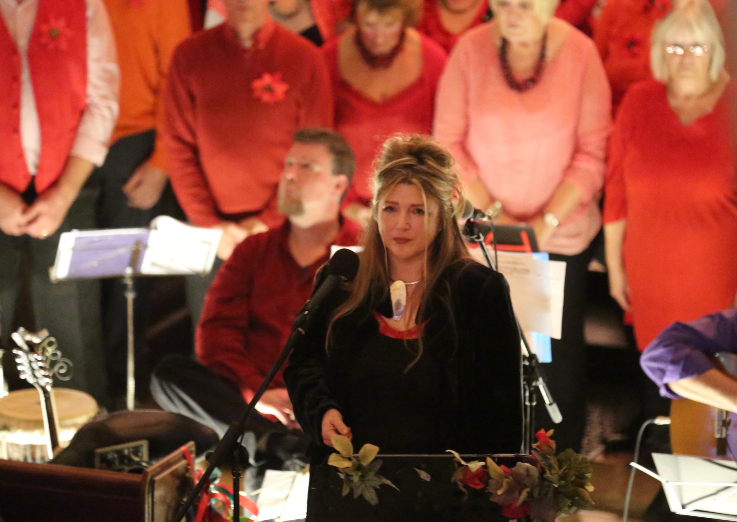 A woman dressed in black stands in front of a choir all dressed in red tones.