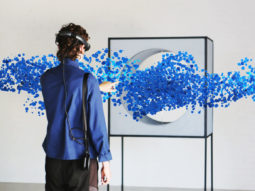A person wearing virtual reality goggles reaches in front of them to touch a stream of blue bubbles flowing through a box with a hole in it.