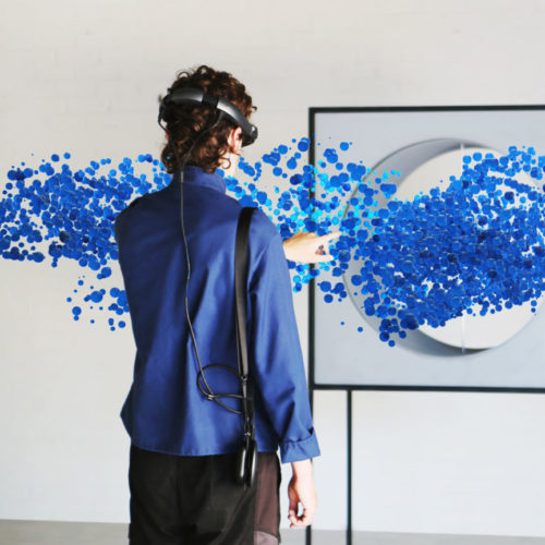 A person wearing virtual reality goggles reaches in front of them to touch a stream of blue bubbles flowing through a box with a hole in it.