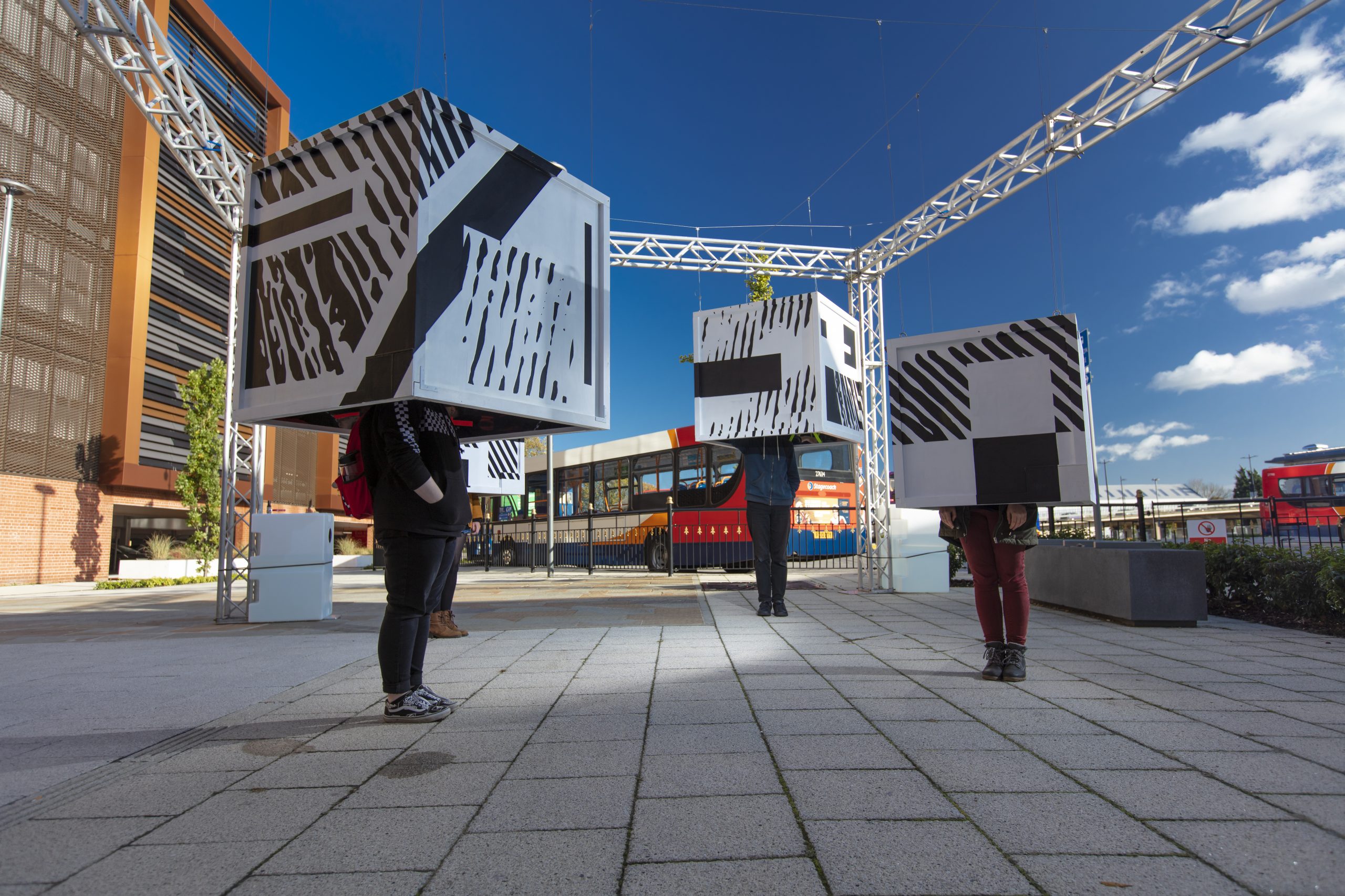 Three large cubes hanging from a frame, three people standing with their heads in them and the rest of their bodies not.